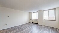 Located at 104 W Franklin St in Richmond, Virginia, this one-bedroom, one-bathroom residence offers a well-organized layout with a clean, modern aesthetic. The living room is the largest area of the home, featuring light gray wood-look flooring, crisp white walls, and multiple windows that bring in an abundance of natural light. A radiator unit provides efficient heating, and the open feel of the space makes it highly versatile for a variety of furniture arrangements. The spacious bedroom continues the same wood-look flooring and neutral color palette, with a window offering views of the surrounding Richmond cityscape. A generously sized closet with a full-width hanging rod provides ample built-in storage. The kitchen, visible in the floorplan overview, is compactly designed with white cabinetry, making efficient use of the available space. The bathroom is finished with large-format beige ceramic wall tiles surrounding a full soaking tub and shower combination, complemented by chrome fixtures for a clean, polished look. Throughout the residence, the consistent use of light tones, modern flooring, and natural light creates a bright, cohesive, and inviting atmosphere well-suited to urban living in the heart of Richmond.