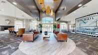 Located at 5151 E Guadalupe Rd in Phoenix, AZ, this impressive single-story clubhouse spans a generous floor area designed to accommodate both social gatherings and everyday relaxation. The main living area immediately commands attention with its dramatic soaring ceilings, anchored by a striking cluster of brushed gold pendant lights that cast a warm, inviting glow throughout the space. Light wood-style flooring flows through the central lounge, transitioning to patterned carpet in the surrounding areas, adding visual depth and texture. Rich brown leather sofas and tufted seating are arranged around a live-edge wood coffee table, creating a welcoming focal point. A bold oversized Air Mail mural serves as a defining artistic statement on one wall, lending the space a distinctive, eclectic character. A rustic reclaimed wood bar counter with bar stool seating and stainless steel appliances provides a functional social hub, bathed in natural light from large arched windows. Additional seating areas include leather barrel chairs positioned before expansive windows overlooking a resort-style pool framed by palm trees. A marble fireplace with an ornamental sculptural accent and a high-top bistro table with industrial stools complete the lounge areas. Throughout, abundant natural light, outdoor views, and curated furnishings create a vibrant yet comfortable communal atmosphere.