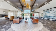 Located at 5151 E Guadalupe Rd in Phoenix, AZ, this impressive single-story clubhouse spans a generous floor area designed to accommodate both social gatherings and everyday relaxation. The main living area immediately commands attention with its dramatic soaring ceilings, anchored by a striking cluster of brushed gold pendant lights that cast a warm, inviting glow throughout the space. Light wood-style flooring flows through the central lounge, transitioning to patterned carpet in the surrounding areas, adding visual depth and texture. Rich brown leather sofas and tufted seating are arranged around a live-edge wood coffee table, creating a welcoming focal point. A bold oversized Air Mail mural serves as a defining artistic statement on one wall, lending the space a distinctive, eclectic character. A rustic reclaimed wood bar counter with bar stool seating and stainless steel appliances provides a functional social hub, bathed in natural light from large arched windows. Additional seating areas include leather barrel chairs positioned before expansive windows overlooking a resort-style pool framed by palm trees. A marble fireplace with an ornamental sculptural accent and a high-top bistro table with industrial stools complete the lounge areas. Throughout, abundant natural light, outdoor views, and curated furnishings create a vibrant yet comfortable communal atmosphere.
