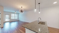 This well-appointed single-floor residence offers a thoughtfully designed open-concept layout that seamlessly connects the kitchen, dining, and living areas. The kitchen serves as the centerpiece of the space, showcasing a large central island topped with granite countertops and an undermount stainless steel sink, complemented by sleek pendant lighting overhead. White shaker-style cabinetry with matte black hardware lines the walls, paired with a full suite of stainless steel appliances including a refrigerator, range, and microwave. Warm hardwood-style flooring flows throughout the main living areas, reflecting natural light that enters through multiple windows. A striking sputnik chandelier anchors the dining area, adding a bold modern design element. The open living area is bright and airy, featuring a ceiling fan and access to natural light through a window-flanked door. The bedroom offers plush carpeting, a ceiling fan, and a window with views of greenery. The bathroom features granite countertops, white cabinetry with matte black accents, a large framed mirror, and direct access to a walk-in closet, completing this clean and contemporary residence.