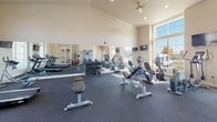 This fitness center offers a spacious, single-floor layout designed to accommodate a comprehensive workout experience. The room is defined by its impressively tall vaulted ceilings, which lend an open, airy feel throughout the space. Recessed lighting and a ceiling fan complement the abundant natural light that streams in through large, floor-to-ceiling windows, providing views of the surrounding neighborhood and keeping the environment bright and inviting.

The flooring throughout is durable dark rubber, ideal for high-traffic fitness use. A full-length mirror spans one wall, visually expanding the room and allowing users to monitor their form during exercise. Mounted flat-screen TVs are positioned at multiple points around the room for entertainment during workouts.

The cardio zone is well-appointed with multiple treadmills, elliptical trainers, and stationary bikes, each equipped with individual digital consoles. A functional cable machine with multiple attachments anchors the strength training area, complemented by a rack of dumbbells and a medicine ball storage tower offering a variety of weights. A dedicated stretching bench rounds out the strength section.

Neutral beige walls and clean white trim contribute to a polished, professional aesthetic. The space is accessible via a glass-paneled door and is clearly marked with emergency exit signage, reflecting thoughtful safety design throughout.