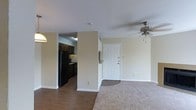 This single-floor space offers an open and functional layout connecting the living area and kitchen. The living room features a combination of hardwood-style flooring and carpeting, with a ceiling fan providing both light and ventilation. Neutral beige walls create a clean, understated backdrop throughout the space. A white paneled entry door serves as the main entryway, providing a welcoming focal point visible from the living area. The kitchen is partially separated by a structural column and wall, offering a degree of definition while maintaining visual connectivity to the main living space. Dark cabinetry lines the kitchen walls, complemented by black appliances including a refrigerator and stove, creating a contrast against the lighter walls. Overhead lighting in the kitchen ensures a well-lit work area. The layout is straightforward and practical, with clear sightlines from the living area to both the kitchen and front entry. The consistent neutral color palette and modest design choices throughout contribute to a cohesive and easy-to-maintain environment suited to everyday living.