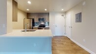 This single-floor residence offers a clean, modern aesthetic with thoughtfully designed living spaces. The open-concept layout connects the living area to a well-equipped kitchen, creating a seamless flow ideal for everyday living. Rich wood-look flooring runs throughout the main living areas, complementing the warm, neutral wall tones.

The kitchen is a standout feature, boasting wood-grain cabinetry, a striking teal subway tile backsplash, white quartz countertops, and a peninsula with an integrated sink. Stainless steel appliances — including a refrigerator, range, and microwave — complete the functional and stylish space. Recessed lighting adds a polished finish overhead.

The living area is generously proportioned, offering ample room for a variety of furniture arrangements. A sliding glass door with louvered shutters allows natural light to pour in while providing access to an outdoor balcony with exterior views.

The bedroom features a neutral palette with wood-look flooring and clean white baseboards, providing a calm and restful environment. The bathroom is finished with a wood-toned floating vanity, white quartz countertop, a large mirror, and a tiled tub-shower combination, offering a contemporary and functional design. Overall, the space presents a cohesive modern style with quality finishes throughout.