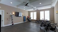 This dedicated fitness room offers a thoughtfully designed workout space that combines functionality with a polished aesthetic. The room features a coffered ceiling with recessed lighting and dual ceiling fans, ensuring proper air circulation during intense exercise sessions. Warm-toned walls are complemented by dark rubberized flooring, which provides both durability and safety for various workout activities. Natural light filters in through multiple windows fitted with white blinds along one wall, creating a bright and energizing atmosphere. A large flat-screen TV is wall-mounted for entertainment during workouts, flanked by decorative wall sconces that add a refined touch. The space is equipped with multiple spinning bikes positioned near the windows, a selection of medicine and stability balls stored on a dedicated rack, and a blue exercise mat leaning against the wall. The open layout allows ample room for movement between equipment, making the space versatile for both cardio and functional training. Overall, the room balances a well-designed interior with practical fitness amenities.
