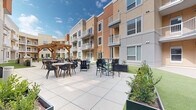 Located on S Homestead Drive in Andover, Kansas, this outdoor courtyard is a well-designed communal amenity space set within a modern multi-story residential building. The courtyard's architecture is defined by a contemporary facade featuring warm earth tones of beige, tan, and burnt orange, with multiple private balconies overlooking the shared grounds below.

The space is divided into two distinct functional zones. A generous paved patio area is furnished with a mix of standard and bar-height dining sets, ideal for socializing and outdoor dining. A wooden pergola anchors one corner of the patio, providing a shaded gathering spot with additional seating. Raised planter beds with manicured shrubs line the transition between the hardscape and lawn areas, adding greenery and structure to the design.

The second zone features a lush, well-maintained artificial turf lawn that provides a clean, versatile surface for recreational activities. Cornhole boards are set up across the green, and lounge chairs are positioned along the perimeter for relaxed outdoor enjoyment. Sliding glass doors from ground-floor units open directly onto the courtyard, creating a seamless indoor-outdoor connection. The open-sky orientation allows abundant natural light to fill the space throughout the day, contributing to a welcoming and active community atmosphere.