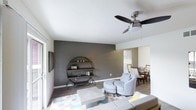 This two-story space showcases a thoughtfully designed interior that blends modern aesthetics with functional living. The main living area immediately draws attention with its bold charcoal accent wall, which serves as a striking backdrop for a distinctive circular shelving unit — a standout architectural and decorative feature that adds both visual interest and display functionality. The living area is anchored by wood-look flooring that runs throughout, complementing the neutral white ceiling equipped with a sleek three-blade ceiling fan with integrated lighting. The open-concept layout allows the living space to flow naturally into the dining area, which benefits from natural light streaming through a window with classic blinds. The dining space accommodates a full table and chair set, making it ideal for everyday meals or entertaining. The color palette throughout is clean and contemporary, with contrasting white walls and dark accent tones creating a balanced, modern ambiance. The two-floor layout offers additional living space beyond what is visible, providing generous room for various lifestyle needs. Overall, the space presents a cohesive, stylish environment with carefully considered design details.