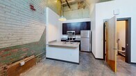 This unit at 144 4th St, Apt 105 in Burlington, NC showcases an industrial-chic aesthetic with exposed brick walls, soaring ceilings with visible wooden beams, and polished concrete floors throughout. The open kitchen is a standout feature, equipped with dark espresso cabinetry, granite countertops, stainless steel appliances—including a refrigerator, range, and microwave—and a peninsula with a double sink. The bathroom continues the design language with a dark vanity, granite countertop, and a clean, well-lit mirror. A stacked washer and dryer unit provides convenient in-unit laundry.
