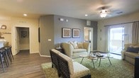 This single-story space showcases a thoughtfully designed living area that serves as the central gathering point. The living room features warm hardwood-style flooring that transitions into plush carpeting, anchored by a green area rug that adds a pop of color and texture. Neutral-toned walls in a calming gray-beige palette create a cohesive and inviting atmosphere throughout.

The space is furnished with a cream-colored loveseat and complementary accent chairs, arranged around a pair of sleek, dark circular coffee tables. Two framed botanical art pieces adorn the gray feature wall, adding visual interest and a natural touch to the décor. A ceiling fan with an integrated light fixture provides both functionality and understated style overhead, while recessed lighting ensures the space is well-illuminated.

A doorway visible from the living area hints at additional rooms beyond, suggesting a practical and connected floor plan. Natural light filters in through draped windows, contributing to the bright and airy feel of the interior. Overall, the space balances modern design elements with warm, comfortable touches, creating an inviting and livable environment.