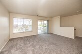 This single-floor residence offers a well-organized, open-concept layout designed for comfortable everyday living. The spacious living area is finished with plush wall-to-wall carpeting and features a textured ceiling, neutral-toned walls, and abundant natural light streaming in through a large picture window and sliding glass door. The sliding door provides direct access to an outdoor area, offering pleasant exterior views and a bright, airy ambiance throughout the space.

The kitchen is thoughtfully designed in a galley configuration, featuring warm honey-toned wood cabinetry lining both sides, white appliances including a refrigerator, electric range, built-in microwave, and dishwasher, all set atop light wood-look flooring. A peninsula with a double sink and a breakfast bar counter seamlessly connects the kitchen to the living area, enhancing the open feel of the space and providing additional prep and seating space. Track lighting illuminates both the kitchen and the adjacent living area transition.

The bathroom is finished with wood-look tile flooring and includes a vanity with warm wood cabinetry, a white countertop, a large mirror, and ample storage. The overall palette throughout the unit is clean and neutral, creating a cohesive and inviting atmosphere that is both practical and easy to personalize.
