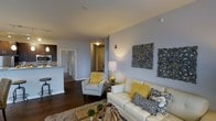 This single-floor residence showcases an open-concept design that connects the living area and kitchen in a cohesive, functional layout. The living room serves as the centerpiece of the space, featuring rich dark hardwood flooring that runs throughout and provides a warm contrast to the soft gray walls. A cream sofa anchors the seating area, complemented by a patterned accent chair, both accented with yellow and gray throw pillows that add a pop of color. Decorative wall panels featuring intricate floral motifs serve as a standout artistic element above the seating area. A floor lamp provides warm ambient lighting, enhancing the inviting atmosphere.

The kitchen, visible from the living area, features a raised breakfast bar with seating, stone countertops, and dark cabinetry, making it both practical and stylish. The open sightlines between the kitchen and living room contribute to a spacious, airy feel. A hallway leading from the living space suggests additional rooms beyond, including what appears to be a bathroom and additional living quarters. The overall ambiance is modern and well-curated, with consistent design elements tying the space together.