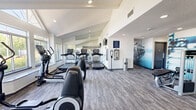 This fitness center is thoughtfully designed across two connected areas, offering a comprehensive workout environment for a wide range of exercise routines. The main cardio zone features vaulted, angled ceilings with recessed lighting and a full-length wall mirror that visually expands the space. Large windows along one wall flood the room with natural light and provide outdoor views, creating an energizing atmosphere. The area is equipped with multiple treadmills, elliptical cross-trainers, and additional cardio machines, all positioned to maximize the use of the generous floor space. Durable patterned carpet tile covers the flooring throughout. Motivational fitness-themed wall murals accent key areas, adding visual interest and energy to the environment. A hand sanitizer station and waste receptacle are conveniently placed for hygiene. The second area transitions into a strength and functional training zone, characterized by soaring ceilings with shiplap detailing and abundant natural light from oversized windows with plantation shutters. This section houses a multi-functional rack stocked with kettlebells, medicine balls, battle ropes, and resistance bands, alongside a dedicated dumbbell rack and a rowing machine. A cable machine and weight bench round out the strength training offerings. The two zones are connected seamlessly, creating a cohesive and fully functional fitness facility.