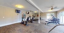 This fitness center offers a spacious, single-floor layout designed to accommodate a well-rounded workout experience. The room is finished with neutral-toned walls, textured carpeted flooring in a multi-tone pattern, and a textured ceiling with flush-mount light fixtures providing consistent overhead illumination. A ceiling fan serves the main workout area, contributing to air circulation during exercise sessions.

The cardio equipment section is prominently featured and includes treadmills, upright and recumbent stationary bikes, an elliptical trainer, and a stair climber, all positioned throughout the open floor plan. A dedicated free weights area includes a full dumbbell rack with a flat bench, ideal for strength training. Multiple wall-mounted flat-screen televisions are positioned for visibility from various workout stations, enhancing the exercise experience.

Large sliding glass doors and windows along one wall allow natural light to fill the space while offering direct views of an outdoor pool area surrounded by manicured landscaping and an iron fence. A kitchenette with wood cabinetry, a sink, and a compact refrigerator provides added convenience. A structural support column is centrally positioned within the space, while colorful wall art adds visual interest throughout.