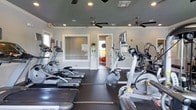 This fitness center offers a comprehensive workout environment designed to support a wide range of exercise routines. The main floor is finished with durable dark rubber flooring, providing a safe and stable surface for all types of training. The cardio area is well-stocked with multiple treadmills, elliptical machines, and stationary bikes, catering to users of varying fitness levels and preferences. Recessed lighting and dual ceiling fans keep the space bright and well-ventilated, creating a comfortable atmosphere during workouts. Large interior windows allow natural light to filter in from adjacent areas, adding an open, airy feel to the room. A doorway leads to a secondary space visible in the background, outfitted with exercise balls in a variety of sizes and colors, ideal for stretching, core training, and group fitness activities. A small storage unit with shelving provides space for accessories and equipment. Informational signage is posted on the walls, offering guidance and safety information for users. The overall layout is thoughtfully organized to maximize usable floor space while maintaining clear pathways between equipment, making this fitness center a functional and inviting place to exercise.