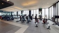 Located on an upper floor of a high-rise building at 1314 Franklin Street in Oakland, CA, this fitness center offers a comprehensive and sophisticated workout environment with sweeping panoramic views of the city and surrounding skyline. The space is designed with a modern aesthetic, featuring a combination of large-format tile and rubber flooring tailored to different workout zones, dark accent columns, recessed and linear pendant lighting, and floor-to-ceiling glass curtain walls that flood the interior with natural light.

The cardio zone lines the perimeter windows, where a row of Life Fitness treadmills and upright bikes are positioned to take full advantage of the elevated city views. Adjacent areas house a variety of additional cardio equipment, including air bikes and rowing machines. The strength training section is well-appointed with a comprehensive free weights area featuring a full dumbbell rack, barbell stations, and multiple cable and resistance machines for both upper and lower body training.

Full-length mirror panels are strategically integrated throughout the space, visually expanding the interior and enhancing the sense of openness. The entry transition from hardwood flooring to the rubber-matted gym floor signals a purposeful separation of spaces. Overall, the fitness center presents a high-end, well-equipped facility that seamlessly blends function with refined architectural detail.