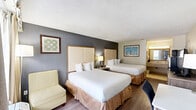 Located at 1400 W Tunnel Blvd in Houma, Louisiana, this hotel room offers a practical and comfortable layout designed to accommodate guests with ease. The spacious main sleeping area features two queen beds dressed in crisp white linens, each with a padded upholstered headboard set against a contrasting accent wall in cool gray tones. Hardwood-style flooring runs throughout the room, lending a clean, modern feel, while a textured ceiling adds classic character overhead. Natural light filters in through a large window dressed with layered curtains, brightening the space. A dedicated work area includes a flat-surface desk with an ergonomic chair, ideal for business travelers, while a wooden dresser with drawers holds a flat-screen television for entertainment. The room features two bathrooms, enhancing convenience for multiple guests. The primary vanity area showcases a wide granite-style countertop with an undermount sink, a framed mirror with overhead lighting, and ample counter space for toiletries. Adjacent to this is a separate water closet with a toilet, towel rack, and wall-mounted amenities. A second full bathroom offers a tub-shower combination with tile surround, a shower curtain, and tile flooring. This room provides a functional and comfortable retreat in the heart of Houma.