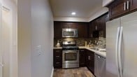 This single-floor space is thoughtfully designed with a contemporary aesthetic and quality finishes throughout. The kitchen serves as a standout feature, showcasing rich, dark espresso-toned cabinetry with sleek bar-pull hardware that contrasts beautifully against light-toned countertops. A full suite of stainless steel appliances includes a gas range, over-the-range microwave, dishwasher, and a single-basin sink, providing a fully functional and modern cooking environment. A multi-toned mosaic tile backsplash adds texture and visual interest to the kitchen walls, complementing the overall color palette. The warm hardwood-style flooring grounds the space with a natural, inviting quality that flows seamlessly through the area. Recessed lighting provides clean, focused illumination, enhancing the kitchen's polished look. The efficient layout of the kitchen maximizes the available space, with upper and lower cabinetry offering ample storage. The neutral wall tones and cohesive design elements create a balanced, welcoming ambiance that is both practical and visually appealing throughout this well-designed space.