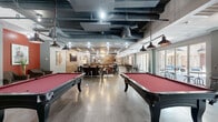 Located at 301 Church Ave in College Station, TX, this generously sized single-floor amenity space offers a dynamic blend of industrial and modern design elements, making it an ideal destination for recreation and socializing. The open-plan layout is anchored by an exposed ceiling with visible ductwork and bold pendant lighting, lending the space an urban, loft-inspired character throughout.

The game area features two full-size pool tables with red felt tops alongside a ping-pong table, providing ample entertainment for groups. Exposed brick walls add warmth and texture, complementing the wood-slat accent walls and mixed flooring that transitions between polished concrete and light hardwood. A built-in wet bar with white subway tile backsplash, bar seating, and shelving serves as a central social hub.

A comfortable lounge zone is anchored by a large leather sectional sofa, area rug, and wooden coffee tables beneath a striking slatted wood ceiling. Additional seating areas with industrial-style chairs and high-top tables are distributed throughout. Large glass sliding doors on multiple sides of the space flood the interior with natural light and provide seamless access to an outdoor courtyard, enhancing the overall sense of openness and connectivity.
