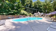 Located at 340 Media Station Rd in Media, PA, this outdoor pool facility offers a well-maintained recreational retreat set against a backdrop of mature trees and natural greenery. The centerpiece is a generously sized kidney-shaped swimming pool with crystal-clear turquoise water, providing ample space for leisure and exercise. The pool features convenient stainless steel entry rails and is surrounded by a broad concrete pool deck that accommodates a variety of seating arrangements, including lounge chairs, standard chairs, and umbrella-shaded tables — ideal for relaxing in the sun or socializing.

A wooden privacy fence lines one side of the pool area, adding a sense of enclosure and seclusion, while a decorative iron fence borders another section of the property. A charming pool house with a distinctive octagonal-roofed turret sits adjacent to the pool, providing functional support facilities for pool operations. Additional outbuildings are also present on the grounds.

The surrounding landscape is rich with tall, leafy trees that provide natural shade and a serene, park-like atmosphere. A multi-story brick residential building is visible in the background, indicating this pool serves as a shared community amenity. The space is well-suited for warm-weather outdoor enjoyment in a tranquil, green setting.