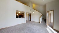 This two-story home is designed with an open and airy feel, anchored by a dramatic vaulted ceiling in the main living area that draws the eye upward and amplifies the sense of space. Plush wall-to-wall carpeting flows throughout the main level, creating a warm and comfortable atmosphere. A kitchen pass-through window connects the cooking space to the living area, promoting an open flow between the two. The kitchen features warm wood cabinetry, pendant lighting, and black appliances, offering a functional and well-appointed setup. A dedicated laundry area is conveniently located on the main floor, housing a stacked washer and dryer unit. A staircase with a wood handrail leads to the second floor, adding to the home's multi-level architectural character. Pendant and flush-mount light fixtures throughout provide consistent, practical illumination. The home also features a private balcony accessible from the upper level, overlooking a well-maintained residential neighborhood with tree-lined sidewalks, manicured lawns, and views of surrounding homes — ideal for enjoying the outdoors in a community setting.