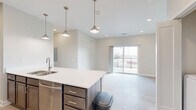 Located at 5504 Tremont Ave in Davenport, Iowa, this well-appointed 2-bedroom, 2-bathroom unit offers a thoughtfully designed single-floor layout. The open-concept kitchen and living area serves as the heart of the home, where a large island with a white quartz countertop and undermount sink anchors the space. Rich, dark-toned cabinetry pairs with stainless steel appliances — including a range, microwave, and refrigerator — and stylish pendant lighting overhead. Light wood-look flooring runs throughout the main living areas, lending a clean, contemporary feel. Sliding glass doors lead to a private balcony, allowing natural light to fill the living space. Both bedrooms feature soft carpet flooring and neutral tones; the second bedroom includes a ceiling fan and a window with outdoor views. The primary bedroom connects to an en-suite bathroom with a dual-sink vanity, white quartz countertops, and a walk-in closet complete with wire shelving. The second full bathroom features a single vanity and a shower/tub combination. A dedicated laundry area and multiple closets throughout provide ample storage. The overall aesthetic is modern and move-in ready, with a bright, neutral palette that creates a clean and welcoming atmosphere.