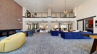This expansive game room and social lounge in Birmingham, Alabama is designed to impress, combining striking architectural elements with versatile entertainment spaces. The interior is anchored by soaring double-height ceilings with an open mezzanine level above, framed by sleek glass railings and dramatic pendant lighting — including geometric chandelier fixtures that command attention throughout the space.

The main lounge area features two distinct flooring zones: a herringbone-patterned wood-look tile and a plush speckled carpet, delineating social gathering areas with bold, colorful furniture including navy sectionals, white sofas, and accent chairs in mustard and red. A textured dark stone accent wall adds depth and visual drama alongside curated wall art.

A built-in wet bar with a dark marble countertop, dark tile backsplash, and vibrant yellow bar stools serves as a central social hub. A media center with an integrated TV and warm wood cabinetry provides an entertainment focal point. Smaller round tables with upholstered chairs create intimate conversation nooks throughout the carpeted lounge area.

A dedicated indoor golf simulator room offers a full hitting bay with turf flooring and a wall-mounted club rack. An additional game area features a billiards table surrounded by large floor-to-ceiling windows that flood the space with natural light and views of the outdoor courtyard.