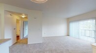This single-floor residential unit is designed with a practical, open layout that connects the main living area to the kitchen. The living room is generously proportioned, featuring neutral wall-to-wall carpeting and crisp white walls that create a bright, clean atmosphere. A sliding glass door fitted with vertical blinds allows natural light to filter in while providing access to the outdoors. HVAC vents are integrated into the ceiling throughout.

The kitchen is positioned adjacent to the living area and is separated by a raised peninsula countertop with a double stainless steel sink, offering both a functional workspace and a natural divider between areas. White cabinetry lines the kitchen walls, complemented by laminate countertops, a white electric range, and a white refrigerator. The kitchen features warm hardwood-style flooring, contrasting with the carpeted living spaces.

The bedroom is a well-sized, carpeted room with access to a dedicated closet and a hallway connection to the rest of the unit. The bathroom includes wood-style flooring, a full tub-shower combination with white tile surround, and a grab bar for added convenience. Overall, the unit maintains a consistent, clean aesthetic with a neutral color palette throughout.