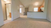 This single-floor space showcases an open-concept layout that seamlessly connects the living area and kitchen. The living space is defined by notably vaulted ceilings that create an airy, expansive feel, with neutral beige walls and wall-to-wall carpet in a warm tan tone contributing to an inviting atmosphere. Natural light filters in through multiple windows, enhancing the brightness throughout.

The kitchen area is positioned to the right, featuring a built-in peninsula with a gray countertop that serves as a natural divider between the cooking and living spaces. Adjacent to the peninsula, wood-toned cabinetry is visible, along with a window above what appears to be a sink area. A track lighting fixture provides dedicated illumination for the kitchen zone.

Toward the rear of the living area, a structural column and partial wall frame a transition point, leading toward what appears to be an entryway with a solid front door. A wall-mounted HVAC vent is also visible, indicating a central climate system. The overall design is clean, functional, and neutral, making the space highly adaptable.