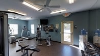 Located at 334 Carolina Forest Blvd in Jacksonville, NC, this fitness center offers a practical and well-equipped space designed to support a range of workout routines. The single-floor facility features a durable rubber flooring surface, ideal for high-traffic exercise use, and is painted in a cool blue-gray tone that lends a calm, focused atmosphere to the room. Natural light filters in through windows and a glass-paneled exit door, keeping the space bright and welcoming. Overhead, fluorescent lighting and a ceiling fan provide consistent illumination and airflow throughout workouts. The cardio equipment lineup includes treadmills and stationary bikes, each fitted with individual monitors for entertainment or tracking during exercise. A wall-mounted flat-screen television serves as a shared viewing option for all users. Informational materials and signage are displayed on the wall, offering guidance for equipment use and facility rules. The space is efficiently laid out to allow comfortable movement between machines, and clearly marked emergency exit access ensures safety. Overall, this fitness center provides a straightforward, functional environment well-suited for residents or members seeking convenient access to essential exercise equipment.