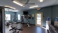 Located at 334 Carolina Forest Blvd in Jacksonville, NC, this fitness center offers a practical and well-equipped space designed to support a range of workout routines. The single-floor facility features a durable rubber flooring surface, ideal for high-traffic exercise use, and is painted in a cool blue-gray tone that lends a calm, focused atmosphere to the room. Natural light filters in through windows and a glass-paneled exit door, keeping the space bright and welcoming. Overhead, fluorescent lighting and a ceiling fan provide consistent illumination and airflow throughout workouts. The cardio equipment lineup includes treadmills and stationary bikes, each fitted with individual monitors for entertainment or tracking during exercise. A wall-mounted flat-screen television serves as a shared viewing option for all users. Informational materials and signage are displayed on the wall, offering guidance for equipment use and facility rules. The space is efficiently laid out to allow comfortable movement between machines, and clearly marked emergency exit access ensures safety. Overall, this fitness center provides a straightforward, functional environment well-suited for residents or members seeking convenient access to essential exercise equipment.