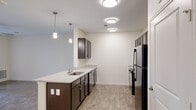 This well-appointed single-floor residence offers a thoughtfully designed layout that balances functionality and modern style. The kitchen serves as a central highlight, featuring rich dark-espresso cabinetry, granite-look countertops, a peninsula island with an undermount sink, and black stainless appliances including a refrigerator, range, and built-in microwave. Pendant lighting above the island adds a contemporary touch, while flush-mount ceiling lights provide ample illumination throughout the space. The kitchen flows seamlessly into the open living area, which is finished with warm wood-look flooring and a ceiling fan overhead. Glass panel doors lead to an outdoor area, allowing natural light to fill the room and offering views of surrounding greenery. The bedrooms are bright and comfortable, each featuring wood-look flooring, large windows with outdoor views, and ceiling fans for added comfort. Several bedrooms include walk-in or reach-in closets with panel-style doors. Neutral wall tones and clean white trim are consistent throughout, creating a cohesive and inviting atmosphere. The overall design prioritizes a bright, open feel with practical finishes suited for everyday living.