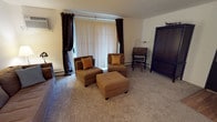 This single-floor residence offers a thoughtfully arranged layout across several distinct living spaces. The living room is generously sized, featuring large sliding glass doors dressed with vertical blinds and dark curtain panels that allow for adjustable natural light. Wall-mounted air conditioning, baseboard heating, and plush carpeting are consistent throughout the space. A secondary seating area provides additional room for relaxation, centered around a sofa beneath framed wall art and flanked by side tables with lamps.

The dining area is defined by a ceiling fan with integrated lighting and accommodates a full-sized table with seating for several guests. The kitchen presents a functional galley-style layout with warm wood cabinetry, laminate countertops, a built-in range, and a full-size refrigerator.

The bedrooms are carpeted and well-lit by natural light from windows with blinds and curtain panels. Each room offers adequate space for standard furnishings. The bathroom features a wide vanity with a large mirror, globe lighting, and tile wainscoting, opening to a carpeted hallway that connects the rooms. Overall, the space maintains a neutral, comfortable ambiance throughout.