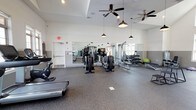 This fitness center offers a spacious, single-level workout environment designed to accommodate a wide range of exercise routines. The open floor plan provides ample room for movement, with durable speckled rubber flooring underfoot that supports both cardio and strength training activities. A selection of Life Fitness treadmills and elliptical machines anchors the cardio section, while a dedicated free weights area features a fully stocked dumbbell rack alongside a stability ball for varied training options. Large wall-mounted mirrors line multiple walls, enhancing the sense of space and allowing users to monitor their form throughout workouts. Natural light filters in through windows along one wall, contributing to a bright and energizing atmosphere. Overhead, dual ceiling fans ensure consistent air circulation throughout the room, complemented by pendant-style industrial lighting fixtures that add a modern aesthetic touch. The white shiplap-style wall paneling, paired with a two-tone wall treatment, gives the space a clean, contemporary look. An emergency exit is clearly marked, and the overall layout prioritizes both functionality and comfort for an effective fitness experience.