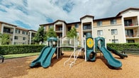 This residential community showcases a warm Mediterranean-inspired architectural design, characterized by a stucco exterior in neutral tones, a clay tile roofline, and private balconies that overlook the landscaped grounds below. The building rises across multiple levels, with generous windows and sliding doors that invite natural light into each unit while offering views of the surrounding outdoor amenities.

A standout feature of this community is the dedicated outdoor playground area, thoughtfully designed for recreation and enjoyment. The play structure includes dual teal slides, a climbing ladder, an interactive spinner panel, and a connecting bridge, all set atop a cushioned wood chip surface that provides a safe environment for active play. The area is framed by well-maintained hedges and mature trees, creating a green, sheltered backdrop that adds to the welcoming atmosphere.

The grounds reflect a commitment to community living, with landscaping that softens the architecture and enhances the overall ambiance. The exterior corridors and balconies contribute to a sense of openness and connectivity throughout the property. Together, the architectural detailing and outdoor recreational spaces make this community a comfortable and engaging environment suited for a variety of residents.