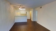 This single-floor space offers an open and functional layout that connects the living area to the kitchen in a seamless, flowing design. Rich, dark hardwood flooring extends throughout the main living area, providing a warm and grounded aesthetic that contrasts with the light, neutral-toned walls.

The kitchen is positioned at the heart of the space, defined by a peninsula-style breakfast bar that serves as a natural divider between the cooking and living zones. White cabinetry lines the kitchen walls, offering ample storage, while an overhead range hood and built-in appliances contribute to a clean, utilitarian finish. A pass-through design enhances the open feel, keeping the space bright and connected.

A ceiling fan with an integrated light fixture is centrally mounted overhead, providing both airflow and ambient lighting to the combined living and kitchen area. Visible HVAC vents are thoughtfully placed to ensure consistent climate control throughout.

Toward the back of the space, a doorway leads to what appears to be a walk-in closet or storage area, adding valuable organizational capacity. The overall ambiance is clean, neutral, and move-in ready, offering a blank canvas that is well-suited for a variety of interior styles and furnishing preferences.