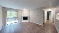 Located in Mill Creek, WA, this single-floor unit at 1108 Mill Creek Blvd delivers a clean, updated interior with a cohesive neutral palette throughout. The living area serves as the heart of the home, featuring durable wood-style flooring, a striking white brick corner fireplace, and a sliding glass door that opens to a private balcony overlooking a lush, wooded backdrop — bringing abundant natural light into the space. The kitchen is well-appointed with crisp white shaker cabinetry, light quartz countertops, a breakfast bar peninsula, and stainless steel appliances including a range and refrigerator. A hallway connects the living and kitchen areas to the private quarters of the home. The bedrooms feature plush carpeting, natural light from windows framing greenery views, and generous closet space — one bedroom notably includes full-width mirrored sliding closet doors. The bathroom is finished with a tub-shower combination, brushed nickel fixtures, and clean white surrounds. A ceiling fan with light fixture is installed in one of the bedrooms, adding comfort year-round. Baseboard heating provides consistent warmth throughout. The overall ambiance is bright, refreshed, and move-in ready, with a serene natural setting just outside.
