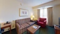 Located at 286 Daniel Webster Highway in Merrimack, New Hampshire, this suite is designed to accommodate both short and extended stays with a thoughtful, functional layout spread across a single floor. The space is divided into distinct living zones, each furnished to support everyday comfort.

The bedroom features a queen bed with a warm wooden headboard, flanked by matching nightstands with table lamps. A dresser with a flat-screen television sits opposite the bed, and large windows dressed in layered curtains allow natural light to fill the room. The signature green patterned carpet runs throughout, grounding the space with a consistent aesthetic.

The living area offers a striped sofa and a plush red armchair arranged around a wooden coffee table, creating a comfortable space for relaxing. Framed artwork on neutral walls adds a quiet decorative touch.

The fully equipped kitchen includes a full-size refrigerator, electric range, microwave, dishwasher, and ample cabinetry — providing everything needed for in-suite meal preparation. Light-toned countertops and warm yellow walls give the kitchen an inviting feel.

The bathroom features a split-style layout, with a separate wet area housing a shower/tub combination and a private toilet, alongside an extended vanity with a large mirror and generous counter space, making the suite practical for multiple guests.