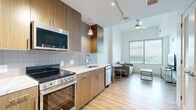 Located at 6919 Main St in Houston's Texas Medical Center district, this compact and efficiently designed one-bedroom, one-bathroom unit maximizes every square foot with a thoughtful, modern layout. Light wood-tone flooring flows consistently throughout, creating a cohesive and clean aesthetic.

The open-plan kitchen and living area serves as the heart of the unit. The kitchen is outfitted with warm wood cabinetry, white quartz countertops, a subway tile backsplash, and a full suite of stainless steel appliances including a range, over-the-range microwave, dishwasher, and refrigerator. Pendant lighting and track lighting provide both task and ambient illumination, while a ceiling fan enhances comfort in the living area. A large window draws in natural light and frames city views.

The bedroom is a bright, well-proportioned retreat featuring an oversized window with sweeping elevated views of the Houston skyline and surrounding cityscape. The space comfortably accommodates a bed and desk area. Adjacent to the bedroom is a dedicated walk-in closet with built-in shelving and hanging rods, offering organized storage.

The bathroom includes a wood-finish vanity with a white countertop, a tub-shower combination with white tile surround, and wood flooring. An in-unit laundry area adds everyday convenience, rounding out this functional and modern urban residence.