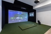 Located in Herriman, Utah, this dedicated indoor golf simulator suite offers a well-appointed, single-floor space designed for an immersive golf experience. The room is cleanly divided into two functional zones: a simulator bay and a lounge area, seamlessly connected by light hardwood flooring that transitions into lush artificial turf.

The simulator bay is the centerpiece of the space, featuring a large projection screen powered by a Foresight Golf system with the XPlay platform, offering access to virtual courses, practice modes, and games. The hitting bay is outfitted with a premium artificial turf mat, providing a realistic feel underfoot. The surrounding walls and ceiling of the simulator area are finished in deep black, minimizing distractions and enhancing the visual impact of the projected display. A ceiling-mounted projector delivers crisp, vivid imagery.

The adjacent lounge area provides a welcoming retreat for players and spectators alike. Light hardwood floors, white walls, and natural light from plantation-shuttered windows create a bright, clean atmosphere. A pair of leather accent chairs with a side table offer comfortable seating, while a bar-height table with stools and a compact beverage station add a social, functional touch. A keypad-secured entry door with glass panels completes the professional, polished design of this thoughtfully equipped golf simulator space.