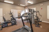 This fitness room offers a dedicated space for a well-rounded exercise experience within a compact, efficiently organized layout. The main exercise area is carpeted and illuminated by fluorescent ceiling fixtures that provide consistent, bright lighting throughout. A large wall-mounted mirror spans one side of the room, visually expanding the space and allowing users to monitor their form during workouts.

The room is equipped with a variety of cardiovascular and strength training machines, including a treadmill, recumbent bike, elliptical trainer, and a multi-station cable weight machine. A wall-mounted television is positioned for convenient viewing during cardio sessions. Shuttered windows along one wall admit natural light and contribute to the room's clean, neutral aesthetic, which features beige-toned walls and white trim.

An adjacent private room, accessible through an interior door, houses a tanning bed and a chair, providing a self-contained space for an additional wellness amenity. This secondary room features its own overhead lighting and carpeted flooring consistent with the main area.

Safety features, including fire extinguishers and clearly marked exit signs, are present throughout. The overall environment is functional, tidy, and purpose-built to support residents' fitness and wellness needs.