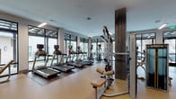 This contemporary fitness center is thoughtfully designed to support a comprehensive workout experience. The space is organized into distinct functional zones, seamlessly connected by an open floor plan with a welcoming entry corridor featuring dark wood wall paneling, decorative wall art, and glass-partitioned rooms that provide visibility throughout.

The cardio zone is generously outfitted with multiple treadmills and elliptical machines, each equipped with individual media screens for entertainment during workouts. Floor-to-ceiling black-framed windows line the perimeter, flooding the space with natural light and offering open views of the surrounding outdoor environment. The combination of rubber sport flooring and hardwood-style flooring throughout provides both comfort and a polished aesthetic.

The strength training area features a robust selection of Matrix cable machines, free weight stations, and multi-function equipment arranged to maximize usable floor space. Rustic dark wood pillar accents add warmth and visual contrast against the clean, light-toned walls and modern linear ceiling lighting. Mirrored wall sections enhance the sense of openness throughout. The overall ambiance is professional yet inviting, striking a balance between a high-performance athletic facility and an upscale, design-forward environment.