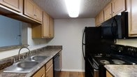 This single-floor residential unit offers a practical and functional layout with a seamless flow between the kitchen and living areas. The kitchen is well-appointed with warm wood cabinetry providing ample upper and lower storage, dark granite-style countertops, and a full suite of black appliances including a refrigerator, electric range, and dishwasher. A double-basin stainless steel sink is positioned beneath overhead cabinetry, and hardwood-style flooring runs throughout the kitchen. A textured ceiling with flush-mounted fluorescent lighting keeps the kitchen bright and well-lit. A peninsular breakfast bar with a dark countertop edge connects the kitchen to the adjoining living space, creating an open and communicative floor plan. The living area features carpet flooring and neutral wall tones that create a warm and inviting ambiance. Louvered closet doors add a classic touch to one wall, while a hallway leads to additional rooms beyond, suggesting a well-organized layout throughout. Wall-mounted climate control and visible ductwork indicate central HVAC throughout the unit. Overall, the space combines functional design with a comfortable residential atmosphere suitable for everyday living.