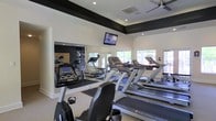 This fitness center is a single-floor facility designed to support a well-rounded workout experience. The space is outfitted with a row of Matrix treadmills and stationary bikes, providing ample cardio options for users of varying fitness levels. Bold dark accent beams frame the upper perimeter of the room, creating a clean visual contrast against the crisp white walls and ceiling. Recessed lighting works in tandem with a ceiling fan to keep the environment bright and comfortable during exercise. A large wall-mounted flat-screen TV is positioned for clear viewing from the cardio equipment, adding an entertainment element to the workout experience. Floor-to-ceiling mirrors line one wall, visually expanding the space and allowing users to monitor their form. Large windows along the exterior wall flood the room with natural light and offer views of the surrounding greenery outside, creating an open, airy atmosphere. The flooring transitions from light-colored material in the open areas to a darker rubberized surface beneath the equipment, providing functionality and safety. Overall, the fitness center combines thoughtful design with practical amenities to deliver a comfortable and motivating environment for daily exercise.