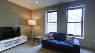 This single-floor urban space offers a clean, modern aesthetic with a practical and welcoming layout. The living area is the focal point of the space, characterized by neutral warm-toned walls and carpeted flooring that create a cozy yet contemporary atmosphere. Two large windows dominate the room, flooding the interior with natural light and framing impressive views of the surrounding city buildings — a standout feature that brings an energetic urban backdrop into the everyday living environment. The windows are fitted with blinds, allowing for flexible light control throughout the day. The space is thoughtfully appointed with understated furnishings, including a deep navy sofa accented with patterned throw pillows, a rustic wooden side table, and a sleek white storage unit. A modern tripod floor lamp adds a stylish accent while providing warm supplemental lighting. Recessed ceiling lighting further enhances the clean, uncluttered look of the space. The overall ambiance is calm and functional, balancing the vibrancy of city life visible through the windows with a subdued, comfortable interior — making it well-suited for both relaxing and everyday living.
