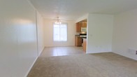 This single-story space showcases an open-concept layout that seamlessly connects the living area to the kitchen and dining space. The living area is finished with plush neutral-toned carpeting, creating a comfortable and inviting atmosphere, while the transition into the dining and kitchen area features light tile flooring for a practical, easy-to-clean surface.

The kitchen is equipped with warm medium-toned wood cabinetry, a built-in microwave, an electric range, and dark countertops that provide a subtle contrast to the light surroundings. The cabinetry offers ample storage space, and the layout allows for efficient use of the cooking and prep areas.

A ceiling fan with a light fixture hangs above the dining area, providing both ventilation and ambient lighting. A large window allows natural light to pour into the space, brightening the open floor plan and enhancing the airy feel. The walls are finished in a clean, neutral white throughout, complemented by a textured ceiling. The overall ambiance is simple, functional, and well-lit, making the space highly versatile.