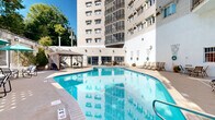 Located at 525 Lexington Pkwy S in Saint Paul, Minnesota, this outdoor amenity area is centered around a generously sized, freeform swimming pool with clear blue water set within a courtyard enclosed by the building's multi-story white brick facade. The pool is surrounded by an expansive concrete deck furnished with lounge chairs and seating areas, providing ample space for relaxation and recreation. A covered gazebo on one side of the pool area offers shaded seating, while umbrella-shaded tables and chairs are positioned along the perimeter for additional outdoor dining and leisure. Decorative wall-mounted artwork accents the bright white exterior walls, adding a touch of character to the courtyard. Lush greenery, potted plants, and mature trees along the boundary walls soften the hardscape and create a pleasant, resort-like atmosphere. Staircase access points are integrated into the courtyard design, connecting the ground-level amenity space to the building above. The courtyard layout takes full advantage of open sky exposure, bathing the entire area in natural sunlight throughout the day. The combination of thoughtful landscaping, varied seating options, and a well-maintained pool makes this outdoor space a standout communal amenity in the heart of Saint Paul.