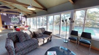 This single-floor space showcases a distinctly mid-century modern aesthetic, highlighted by dramatic vaulted ceilings with exposed wooden beams that add architectural character and a sense of openness. The ceiling features a textured finish in a muted tone, complementing the warm tones of the structural beams overhead.

The living area is generously proportioned, anchored by a patterned sofa and a sleek, dark glass-topped coffee table. A retro-style multi-arm floor lamp adds a sculptural element to the space, while a mid-century accent chair provides additional seating. The carpeted flooring contributes a casual, comfortable feel throughout.

The most striking feature is the expansive wall of floor-to-ceiling sliding glass doors and windows, which floods the interior with abundant natural light and offers unobstructed views of an outdoor swimming pool and surrounding mature trees. This seamless indoor-outdoor connection creates a resort-like ambiance. Additional seating and a dining area are visible toward the rear of the space. A pendant barn-style light fixture overhead reinforces the space's clean, functional design language.