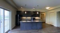 This contemporary single-floor space showcases a thoughtfully designed open-concept layout that seamlessly blends style and functionality. The kitchen serves as the centerpiece, featuring rich dark wood cabinetry that extends to the ceiling, providing generous storage throughout. A large central island with a striking dark wood finish and light granite countertop offers ample workspace and serves as a natural gathering point. The island is equipped with a deep sink and is illuminated by elegant pendant lights that hang at just the right height, adding warmth and visual interest. Premium black stainless steel appliances, including a refrigerator, microwave, and range, complement the sophisticated color palette. A tasteful tile backsplash in a neutral tone adds texture between the countertops and upper cabinets. Wide-plank hardwood flooring flows throughout, grounding the space with a warm yet modern aesthetic. The open floor plan extends beyond the kitchen, revealing a bright and airy living area that benefits from the connected layout. Recessed lighting and ceiling-mounted fixtures provide consistent illumination throughout. The overall ambiance is sleek, modern, and inviting, with a cohesive dark-toned design language that conveys both elegance and practicality.