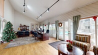 This generously sized single-floor space offers a well-organized layout that combines functional office use with comfortable living amenities. The office area is a standout feature, characterized by vaulted, angled ceilings clad in white shiplap-style paneling and illuminated by a track lighting system. Large windows dressed with sage green curtains and horizontal blinds line the walls, providing direct views of an outdoor pool area and allowing plentiful natural light to fill the room. French doors offer direct access to the exterior. Light hardwood flooring runs throughout, complementing the warm wood tones of the executive desk and conference-style seating arrangement. A relaxed seating area with a round wood coffee table and striped upholstered chairs is positioned near the entrance, creating a welcoming reception-style zone. The kitchen and living room area features a peninsula breakfast bar with dark leather bar stools, wood-toned cabinetry, dark granite countertops, and standard appliances, with vaulted ceilings adding a sense of openness. A carpeted staircase with natural light from an upper window connects to additional levels. The overall ambiance is warm and professional, blending practical workspace functionality with comfortable, welcoming design elements.