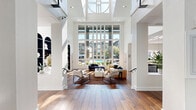 Located at Asbury Square in Dunwoody, Georgia, this impressive single-floor community space is defined by its dramatic architectural design and bright, open atmosphere. The layout flows seamlessly from a welcoming entry hallway — finished with warm hardwood flooring and crisp white walls — into a grand main living area that commands attention with its soaring double-height ceilings supported by bold white structural columns.

The centerpiece of the space is a breathtaking floor-to-ceiling window wall that floods the interior with natural light and frames sweeping views of the surrounding community. The lounge area is thoughtfully arranged with contemporary seating, including plush sofas and modern accent chairs, anchored by a textured area rug that adds warmth to the expansive hardwood floors.

A built-in arched bookcase along one wall adds a refined architectural detail, while an upper-level mezzanine with open railings contributes to the sense of vertical grandeur. The integrated kitchen area features stone countertops, dark cabinetry, stainless steel appliances, and stylish pendant lighting, opening directly to the main space for a cohesive, connected feel. Lush potted greenery throughout the space adds a fresh, organic touch, complementing the clean modern aesthetic and creating an inviting, well-appointed environment.