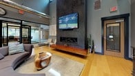 This striking single-floor commercial space blends industrial and contemporary design elements to create a sophisticated and welcoming environment. Soaring vaulted ceilings with exposed ductwork and warm wood beam accents lend an open, loft-like character throughout. Light bamboo hardwood flooring runs continuously across the space, contributing to a bright and cohesive aesthetic.

The central lounge area is anchored by a dramatic floor-to-ceiling textured feature wall finished in dark concrete-style material, housing a sleek linear fireplace and a large wall-mounted flat-screen television. A floating rich wood media console completes this focal point. Unique organic furnishings, including a sculptural live-edge coffee table and curved sectional sofa, add artistic flair to the seating area.

A dedicated dining and bar-height table zone sits beneath gallery-style artwork, bathed in natural light from large windows offering views of the surrounding landscape. Adjacent to this is a well-appointed kitchenette area featuring dark wood cabinetry, a striking marble-topped island with integrated ambient LED lighting, and pendant lighting overhead.

Glass-partitioned rooms with dark metal framing provide semi-private spaces visible from the main lounge, enhancing the open yet functional layout. Recessed lighting and carefully placed floor lamps ensure a warm, inviting ambiance throughout this thoughtfully designed space.